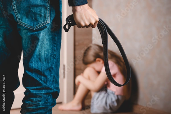 Fototapeta A man with a belt in his hand threatens a little girl. A little girl sits in a corner and hides her face . The concept of domestic violence against a child.