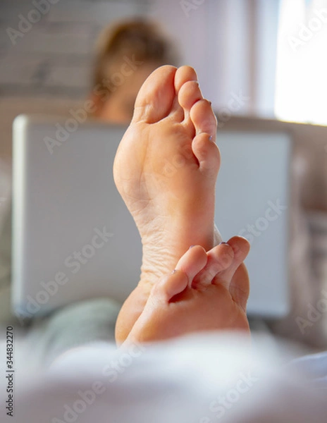 Fototapeta Focus on the feet in the foreground. A teenage girl is lying on a bed with a laptop.