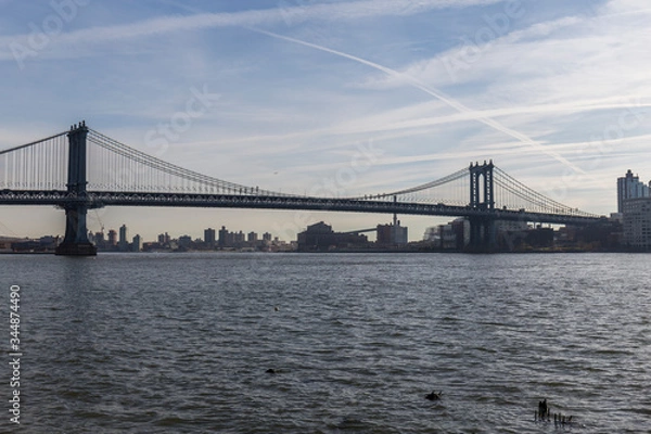 Fototapeta Brooklyn bridge seen from afar, side of the bridge, you can see the dark water river and the blue sky, behind the bridge city buildings