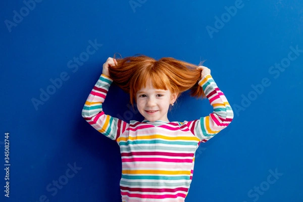Fototapeta little girl with red hair in a multicolored smile stands on a blue background