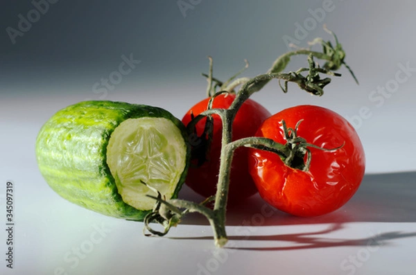 Obraz Wrinkled tomatoes and a half of cucumber lie on a table with a ray of light. Gray background. Hard shadows. Selective focus. Dimmed background.