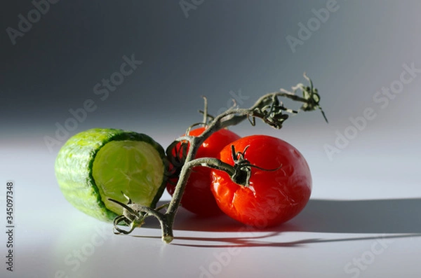 Obraz Wrinkled tomatoes and a half of cucumber lie on a table with a ray of light. Gray background. Hard shadows. Selective focus. Dimmed background.