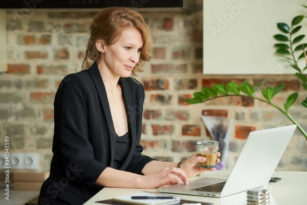 Obraz A happy woman working remotely on a laptop in a kitchen. A blond lady with a coffee discussing a project with colleagues at a video conference at home. A teacher preparing for an online lecture