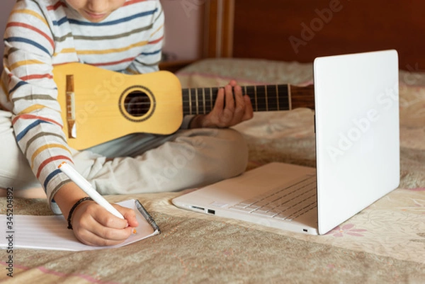 Fototapeta The boy try to learning the timple lesson at home from the internet with his laptop. He writes a new song with chords in his notebook