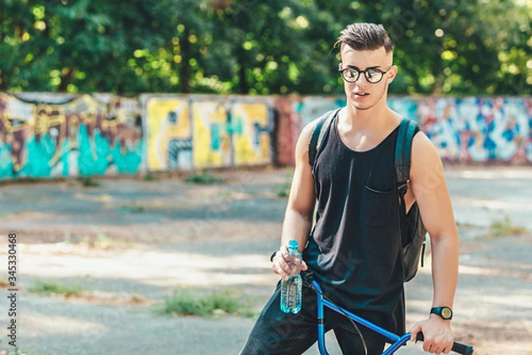Fototapeta Young stylish man with a bicycle and a bottle of water poses in the park. graffiti wall background