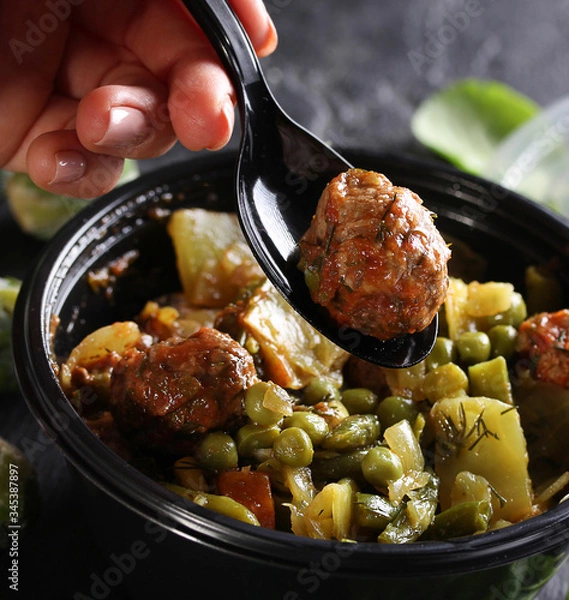 Fototapeta Healthy green food. Stewed cabbage, potatoes, peas and meatballs in a black plastic bowl with a spoon and hand. Green brussels sprouts, spinach on a black background. Background image, copy space