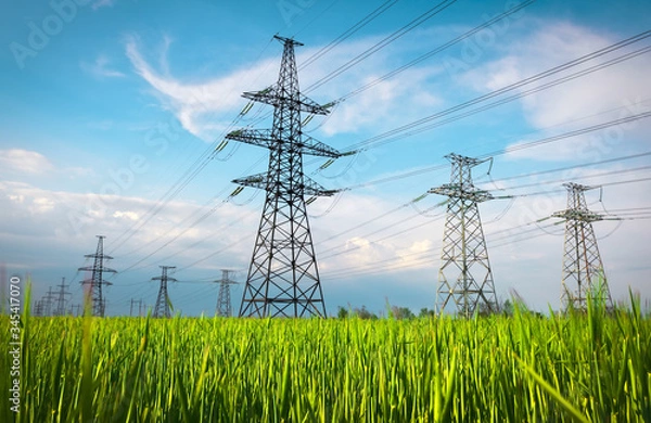 Obraz High voltage lines and power pylons in a flat and green agricultural landscape on a sunny day with clouds in the blue sky. Cloudy and rainy. Wheat is growing
