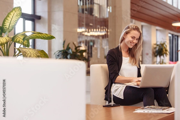Obraz Laughing young beautiful blonde woman is sitting in lobby of hotel and waiting for check-in checking mail and social networks using a laptop and high-speed Internet. Successful beautiful woman concept