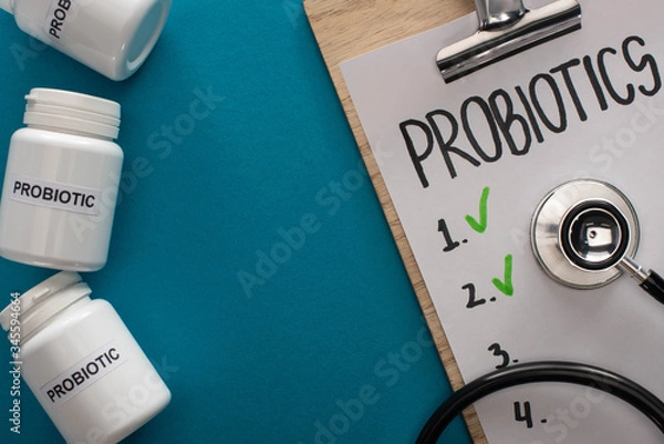 Fototapeta top view of clipboard with probiotics empty list with stethoscope near containers with pills on blue background