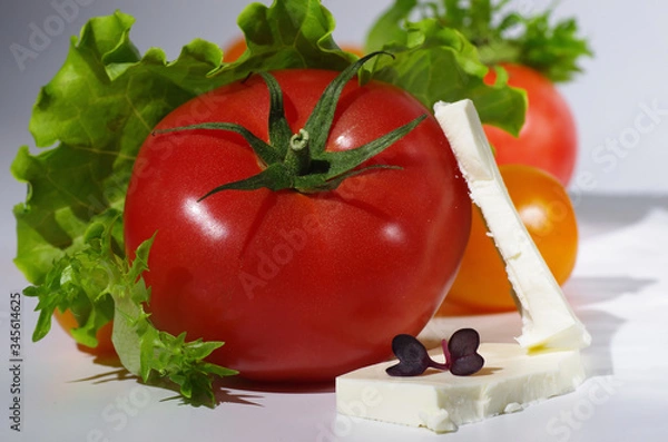 Obraz Pink tomato and sliced feta cheese lies among yellow cherry tomatoes, lettuce leaves and microgreen. Close-up. Gray background. Selective focus.