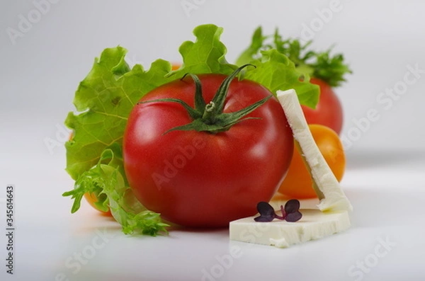 Obraz Pink tomato and sliced feta cheese lies among yellow cherry tomatoes, lettuce leaves and microgreen. Close-up. Gray background. Selective focus.
