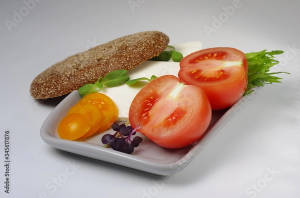Obraz Sliced pink and yellow tomatoes and feta cheese on a plate with a piece of brown bread. Gray background. Selective focus.