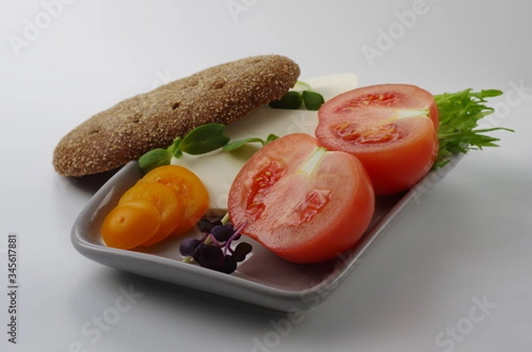 Obraz Sliced pink and yellow tomatoes and feta cheese on a plate with a piece of brown bread. Gray background. Selective focus.