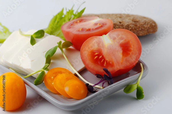 Obraz Sliced pink and yellow tomatoes and feta cheese on a plate with a piece of brown bread. Gray background. Selective focus.