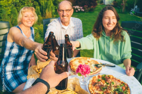Obraz POV image of people toasting with beer bottles at multi generation family backyard party (focus on first bottle)