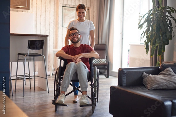 Fototapeta Woman pushing wheelchair with disabled man around the house