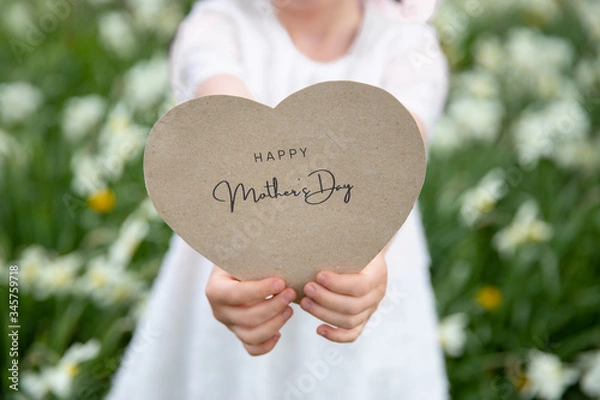 Obraz Happy Mother's Day written on a heart-shaped card held by a girl on flower and grass background