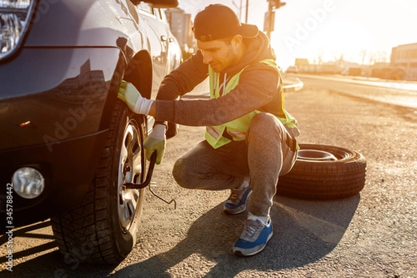 Fototapeta Worker changes a broken wheel of a car. The driver should replace the old wheel with a spare. Man changing wheel after a car breakdown. Transportation, traveling concept