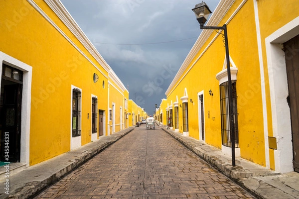 Obraz Beautiful perspective yellow street of the famous city of Izamal in Mexico. A snow-white carriage rides against the background of yellow houses and clouds