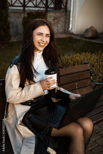 Obraz close portrait of a girl with a laptop sitting on a bench with a cup of coffee. Student with laptop studying in the street.
