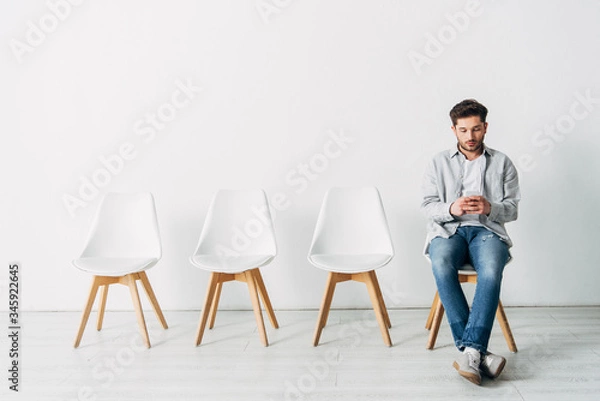Fototapeta Handsome employee using smartphone while sitting on chair in office