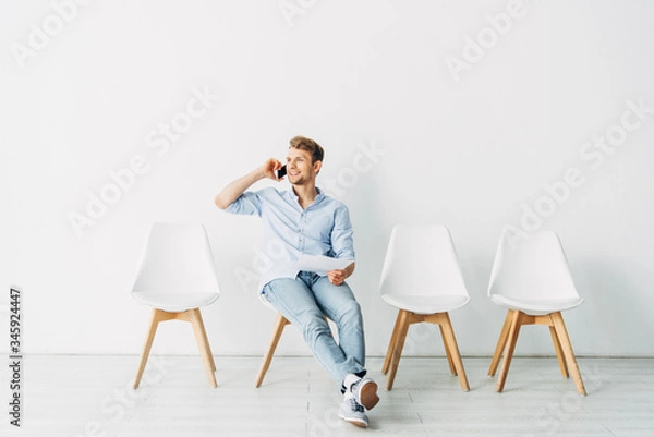 Fototapeta Smiling man talking on smartphone and holding resume in office