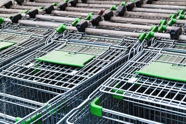 Obraz Empty metal carts on wheels for food or large size things. A pattern of inserted in each other steel grocery carts, standing under an plastic awning in a shopping center parking lot
