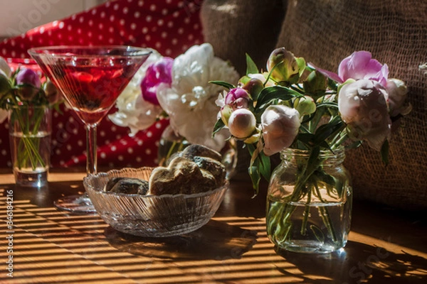 Fototapeta Solemn rustic still life with glass of red drink with cakes in a bowl surrounded by white flowers on textile background on wooden table. Diagonal striped shadow of the blinds on bright sunny day