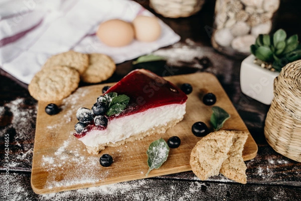 Obraz Slice of blueberry cheesecake on a wood counter, with some berries, biscuits, eggs, green leaves, and red lines on the side.
