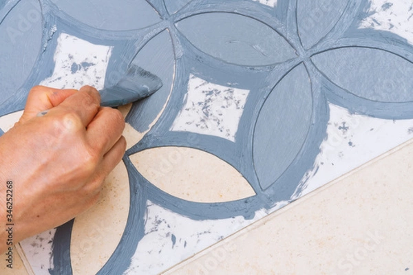 Fototapeta Stay at home and home improvement concept: Close up and top view of a hand holding a brush is painting a decorative template on the floor tiles into gray by using a vintage pattern stencil