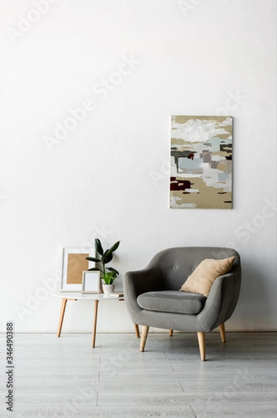 Fototapeta grey armchair near coffee table with frames and green plants near painting on wall in modern living room
