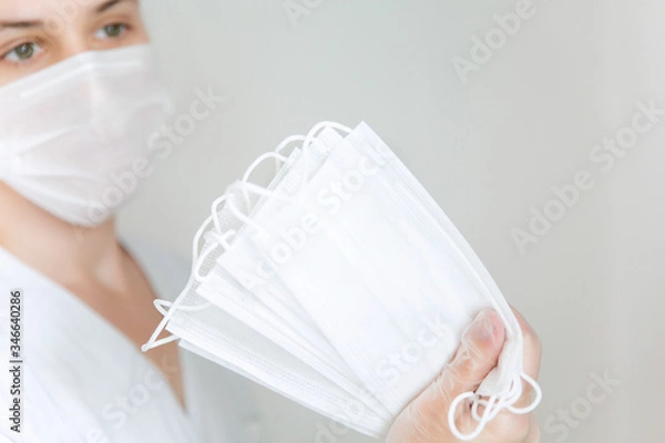 Fototapeta White protective medical masks in hand in the foreground ,a woman with a mask in the background on the left.