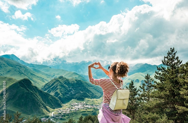 Fototapeta Young female making hands in heart shape sign on the peak of mountains. View in the top of mountain on a hot summer day. Lifestyle concept. Back view. mountain landscape. Freedom concept.