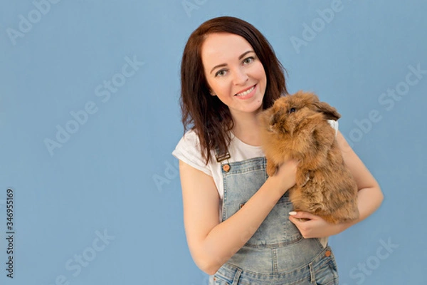 Obraz Portrait of a happy, cute girl 30-33 years old on an isolated blue background. A woman holds a rabbit in her hands.