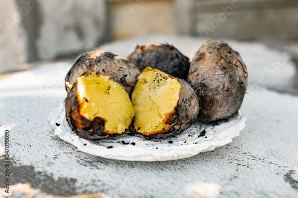 Fototapeta Baked in the fire potatoes in the skin according to the village recipe and method, directly in the ashes. Charred, fragrant tubers, a hot dish on a white stone plate