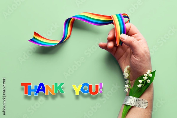 Fototapeta Rainbow ribbon in woman's hand. Chamomile flowers and grass bouquet attached to wrist with medical patch. Thank you doctors and nurses! Creative flat lay, top view on green mint background.