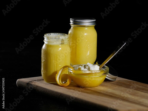 Obraz Homemade lemon curd served in a glass bowl with whipped cream and a lemon peal slice on wood cutting board agains a black background.