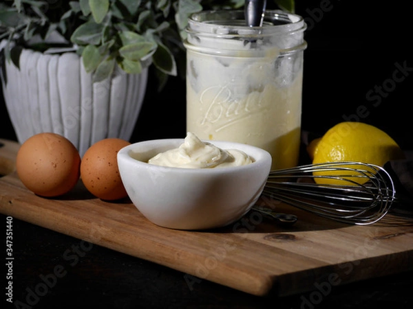 Obraz White bowl of homemade mayonnaise displayed with ingredients including brown eggs and a fresh lemon in a jar on a wood cutting board against a black background. 