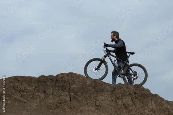 Fototapeta Sports brutal bearded guy on a modern mountain bike. Beautiful view from the mountain. Panoramic view for banner.