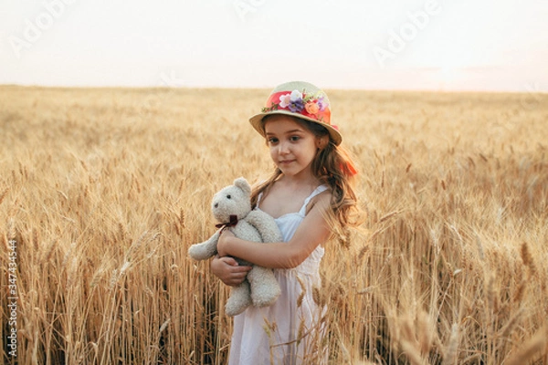 Fototapeta girl in a white dress and a hat with a flower stands in an embrace with an ivy bear