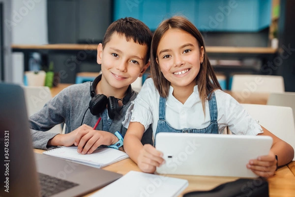 Fototapeta Two happy small friends writing homework , using smart digital tablet at modern blue classroom.