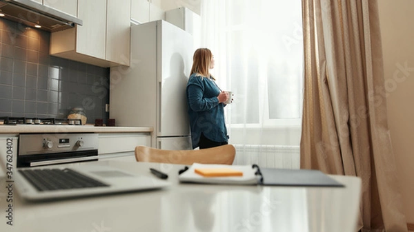 Fototapeta Advantages of working from home. Young woman in casual clothes drinking coffee or tea, looking in the window while standing in the kitchen at home