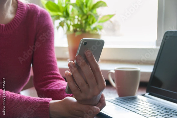 Fototapeta Close up of a woman using smartphne on kitchen table with coffee Mug and laptopNewspaper and Checking Cell Phone Messages on Relaxing Morning at Home
