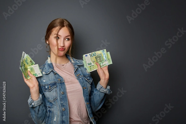 Obraz Portrait of a natural and authentic beautiful young woman, teenager with many 100 Euro banknotes in her hand is happy about the money, in front of a grey wall
