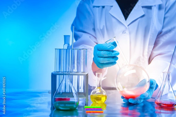 Fototapeta Chemical laboratory. The concept is to conduct chemical experiments. Lab technician's hands with a flask close-up. Chemical dishes and reagents on the table. Analysis of synthetic substances.