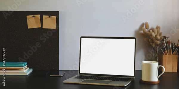Fototapeta Photo of white blank screen computer laptop putting on black working desk and surrounded by stack of notebook, smartphone, pencil holder, wild grass in vase and coffee cup.