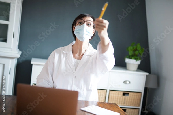 Fototapeta Frau arbeitet in Arztkittel und Mundschutz im Homeoffice oder Praxis zuhause.	
