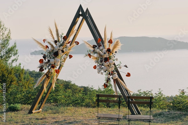 Fototapeta Original wedding arch on top of the mountain with white and red flowers at sunset. Romantic ceremony in the style of boho