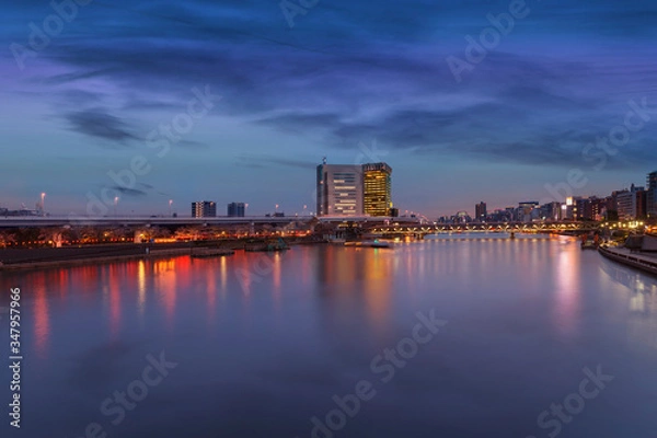 Fototapeta Sumida riverside apartments with skyscrapers of Asahi Beer Headquarter and Bunkyo City Hall at sunset with cherry blossoms trees illuminated.