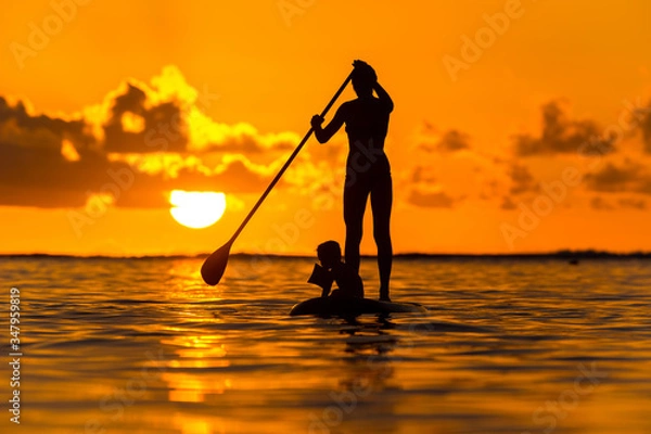 Obraz Young mother with a small child ride a S.U.P. (paddle) board in the Indian Ocean on the background of an incredible sunset